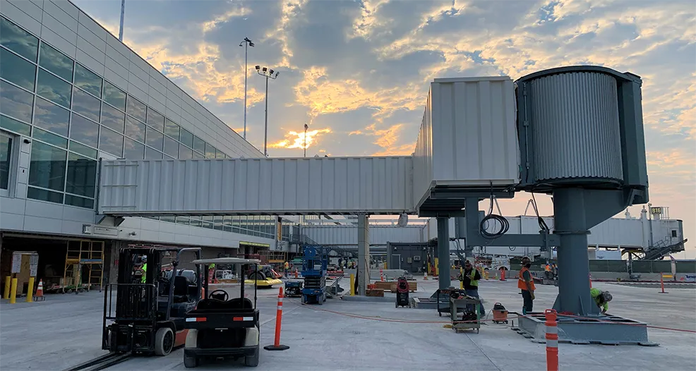 Passenger boarding bridge being installed on the airport ramp at dusk, with crew and lift equipment